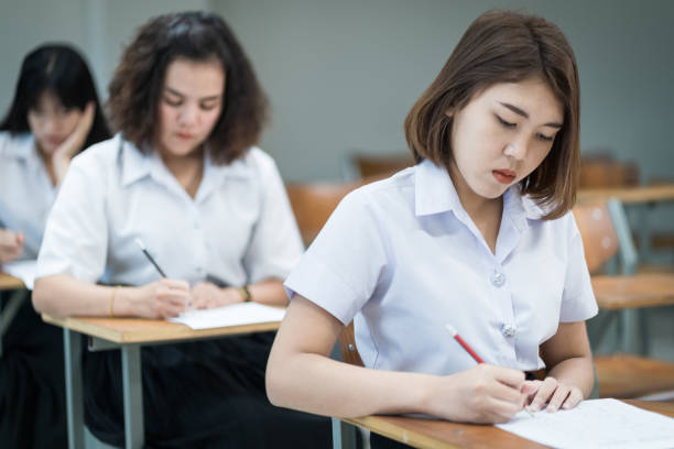Selective focus of the teenage college students sit on lecture chairs do final examination and write on examination paper answer sheets in the classroom. University students in uniform in classroom. stock photo
