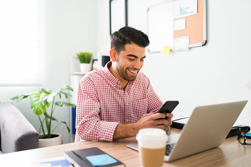 Smiling young man is sitting at his desk while looking at his smartphone. Young guy working as a salesperson is happy to be closing a deal with a client
