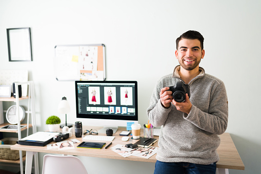 Smiling photographer in his 20s holding a proffesional camera and leaning into his work desk while checking his new photos