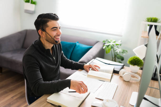 Young guy is very happy to be working remotely as a translator Good-looking young man sitting at his desk and wearing a headset. Smiling guy working as a translator or interpreter with a notebook and books working at home stock pictures, royalty-free photos & images