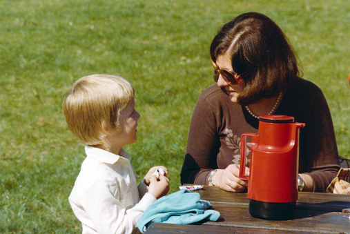 Vintage 1979 color image, young mother with sunglasses  talking with and looking at her son having a roadside picnic with retro red thermos.