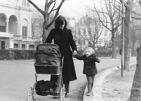 1976 vintage monochrome image of a mother with a long black coat walking hand in hand with her daughter and pushing a classic pram in Vondelpark, Amsterdam, The Netherlands.