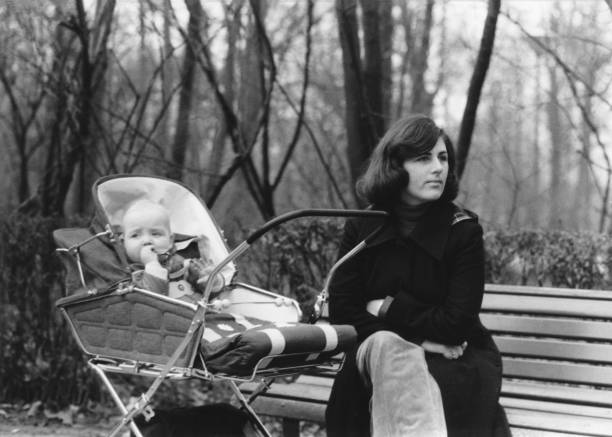 Vintage mother with baby on a bench in the Vondelpark in Amsterdam, The Netherlands 1976 vintage monochrome image of a mother with a long black coat sitting on a wooden bench in the Vondelpark in Amsterdam with her son in a classic pram. 1970s woman stock pictures, royalty-free photos & images