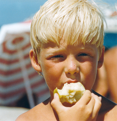 Vintage colorful retro 1979 image, close-up portrait of a young boy with blond hair and beach umbrella background eating an apple.