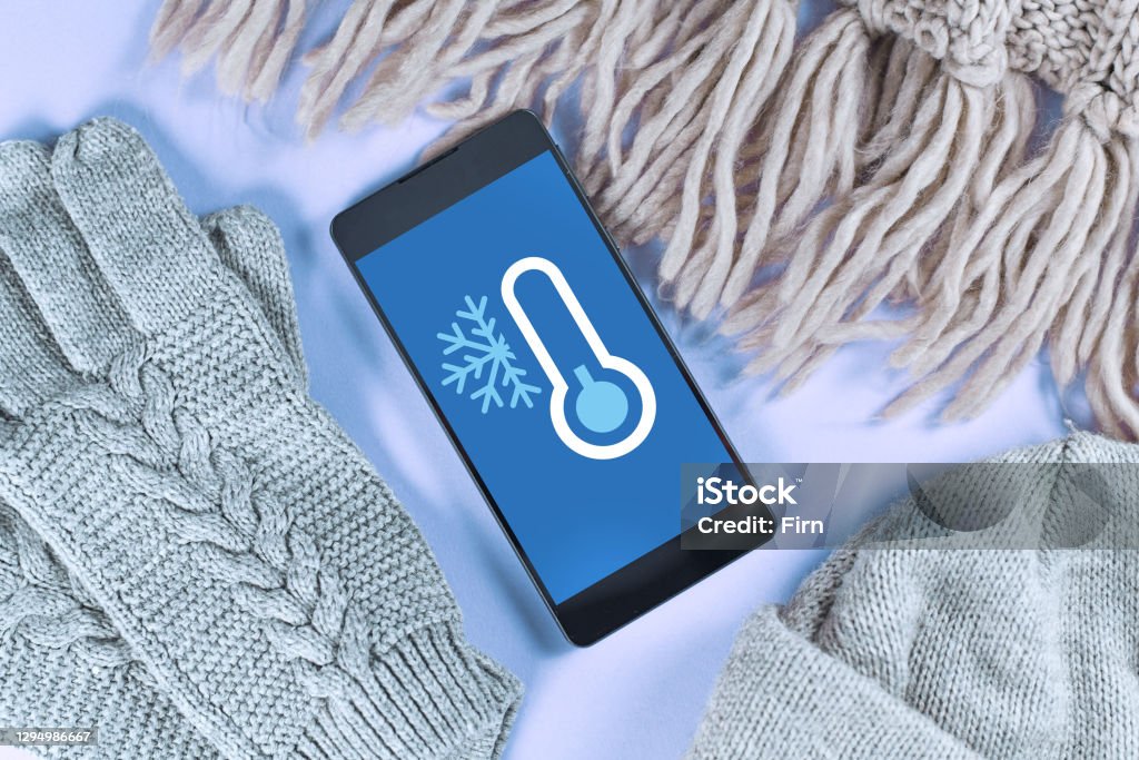 Concept for cold temperatures with snow and minus degrees with mobile phone showing weather forecast surrounded by warm clothes Concept for cold temperatures with snow and minus degrees with mobile phone showing weather forecast surrounded by warm winter clothes like scarf, hat and gloves Cold Temperature Stock Photo