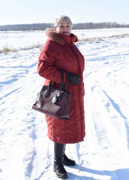 donna anziana russa in camice rosso che guarda la telecamera contro il campo innevato. inverno - russian ethnicity cold relaxation nature foto e immagini stock