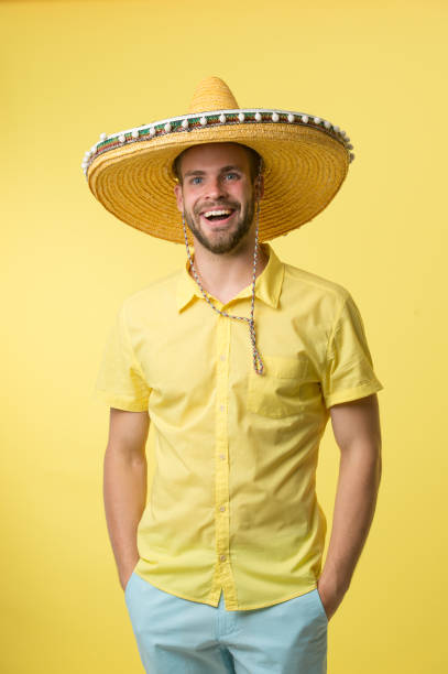 Man on smiling face posing in sombrero hat with hands in pockets, yellow background. Guy with bristle looks festive in sombrero. Fest and holiday concept. Man in festive mood going to celebrate.