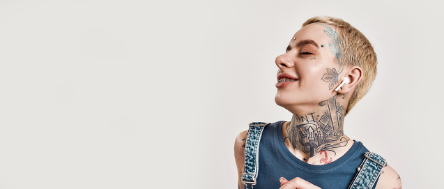 A white girl pierced with tattoos wearing denim overall standing and listening to music with her wireless earphones eyes closed smiling and laughing a bit on a white background