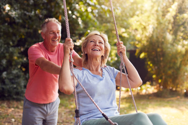 пенсионеры пара весело с мужчиной толкая женщину на сад качели - senior adult senior couple couple summer стоковые фото и изображения