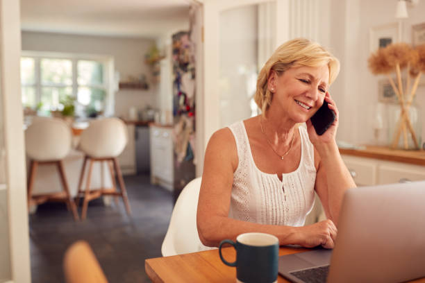 istock Retired Woman On Phone At Home In Kitchen Using Laptop 1295001303