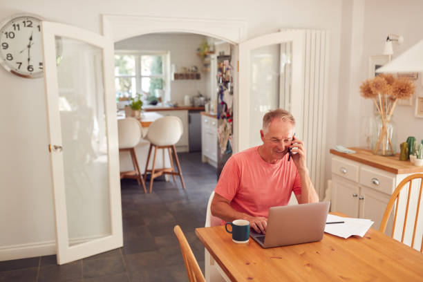istock Retired Man On Phone At Home In Kitchen Using Laptop 1295001734