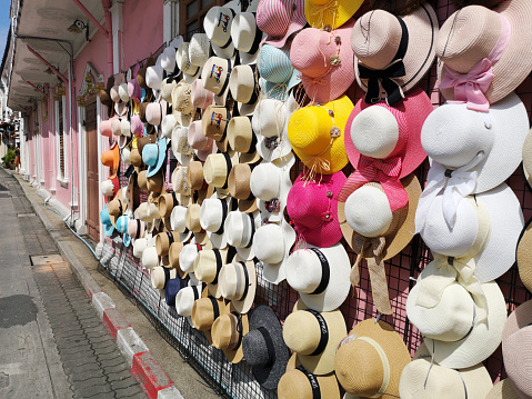 Phuket, Thailand - October 7, 2018: One of the curbside shops in the local weekend market. A variety of fashionable and colorful hats are displaying on the pink building wall for sale. Sideview of store stand with a row of hats in different colors and designs.