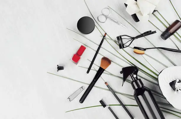 Spring stylish glossy black cosmetic products, mackup accessories with red nail polish, green blades of grass on light white wooden background, border, copy space.