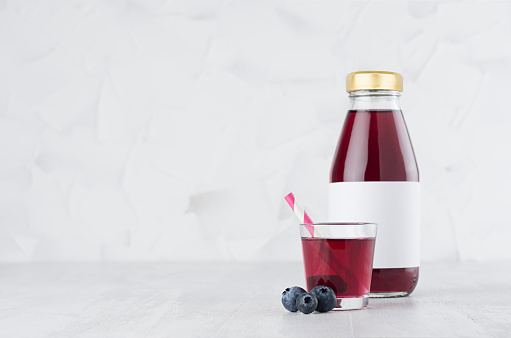 Fresh purple blueberry juice in glass bottle with blank label mock up with glass, straw, berries in soft light white interior on wood table with copy space.