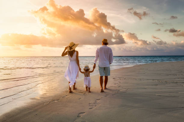 eine familie geht bei sonnenuntergang hand in hand einen tropischen paradiesstrand hinunter - beach stock-fotos und bilder