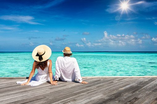 A beautiful couple in white summer clothing sits on a wooden pier and enjoys the view to the turquoise ocean of the tropical Maldives islands