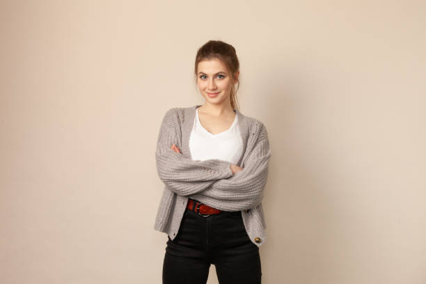 Studio portrait of 18 year old woman with brown hair Studio portrait of an attractive 18 year old woman with brown ponytail hair in a gray cardigan sweater and black pants on a beige background three quarter length stock pictures, royalty-free photos & images