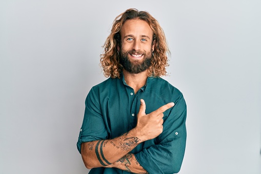 Hombre guapo con barba y pelo largo usando ropa casual sonriendo alegre señalando con la mano y el dedo hacia un lado photo