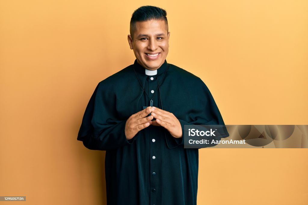 Young latin priest man standing over yellow background hands together and fingers crossed smiling relaxed and cheerful. success and optimistic Priest Stock Photo