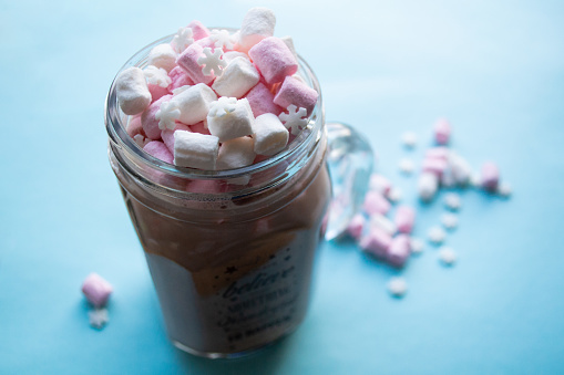 Glass mug with hot cocoa or coffee with milk, sprinkled with white and pink marshmallows and sugary snowflakes on top, on a blue background.