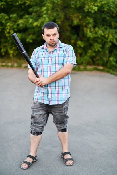 um homem violento de camisa e shorts parado na rua na calçada com um bastão na mão contra o fundo da vegetação - bat weapon baseball mob - fotografias e filmes do acervo