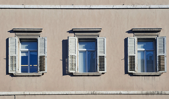 Three classic italian windows on the beige color wall facade with open wooden shabby white shutters