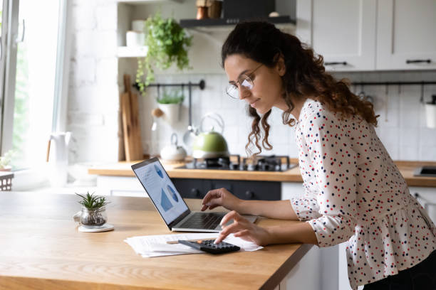 Serious woman wearing glasses calculating finances, working with project statistics Serious young woman wearing glasses calculating finances, household expenses, confident businesswoman working with project statistics, using laptop and calculator, standing in kitchen at home savings account stock pictures, royalty-free photos & images