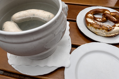two Bavarian white sausages in hot water, a pretzel on a plate and an empty plate