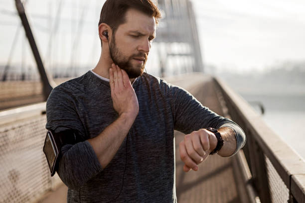 Young athlete runner in headphones looks at a smart watch holding carotid with fingers Young athlete runner in headphones looks at a smart watch taking pulse stock pictures, royalty-free photos & images