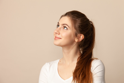 Close-up studio portrait of attractive 18 year old woman with brown ponytail hair in a white t-shirt on a beige background