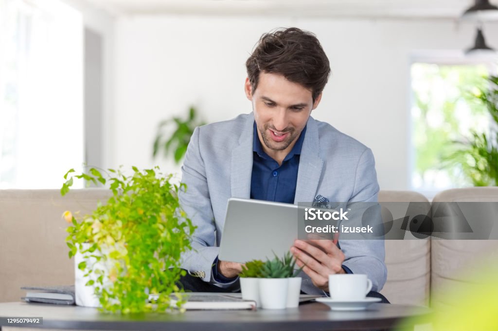 Businessman working in the eco-friendly green office Mid adult men wearing grey jacket sitting on sofa in the creative workplace and using digital tablet. Men Stock Photo