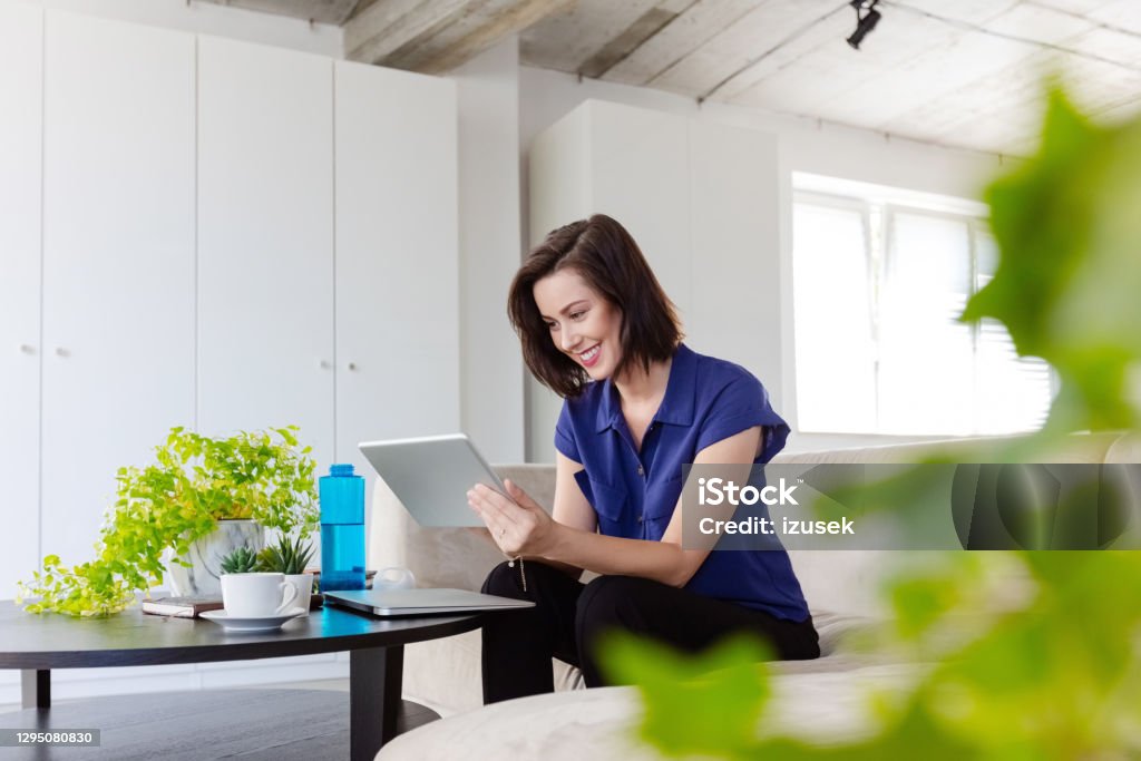 Businesswoman working at home Mid adult women sitting on sofa in the creative eco-friendly workplace and using digital tablet during video call. Office Stock Photo