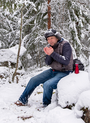 A man, a Caucasian, is frozen and drinking hot coffee in a snowy forest. Winter hiking. The man is resting and enjoying the quiet.