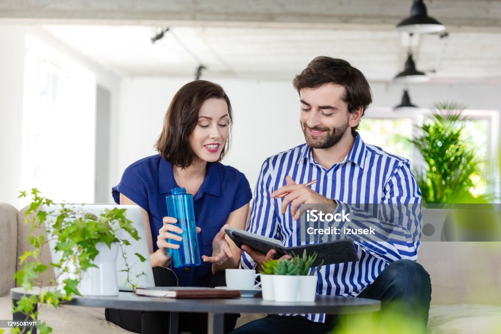 Coworkers working in the eco-friendly green office Mid adult women and man sitting on sofa in the creative workplace and working together, discussing agenda. Woman holding water bottle in hand. Men Stock Photo