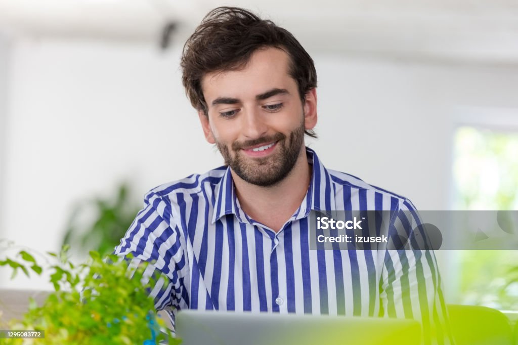 Smiling man using laptop in a green office Mid adult men wearing striped shirt sitting in the creative workplace, using laptop during video conference. 30-39 Years Stock Photo