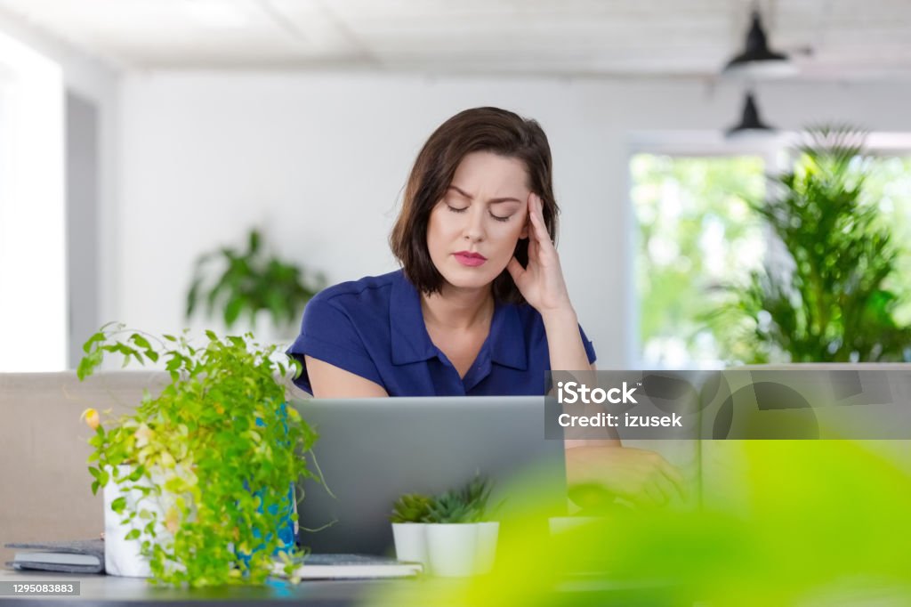 Tired woman working on laptop at home Mid adult women sitting on sofa in the creative eco-friendly workplace and using laptop during video call, suffering from headache. Green Color Stock Photo