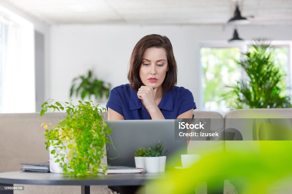 Worried woman working on laptop at home Mid adult women sitting on sofa in the creative eco-friendly workplace and using laptop during video call. Blue Stock Photo