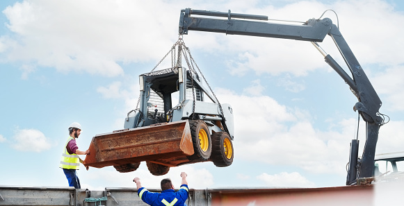 Unloading of skid steer loader by crane at the construction site with workers guiding the driver