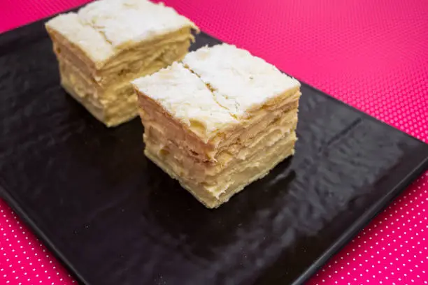 Cream pie with layers of puff pastry in a ceramic plate and a pink background