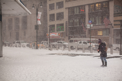 A young woman with a phone in her hands stands in front of the entrance to Penn Station. Everything around is covered with snow. No snow only on street ventilation bars. Snow continues to fall and snowflakes are visible flying in the air. Cars drive along the roadway completely covered by snow. Winter storm in progress and snow gathers in drifts. Tall city buildings of middle Manhattan are visible on a blurred background. Very bad weather. Mid day. January 04, 2018. NYC. Midtown Manhattan. New York. USA