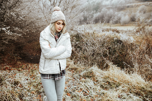 Young blonde female wearing knit hat, white jacket and denim jeans taking a walk in a forest.