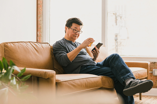 While sitting on a sofa in his home, a senior man concentrates while reading the label on a prescription medication. He is using the pharmacy's mobile app on his smartphone to refill the prescription.