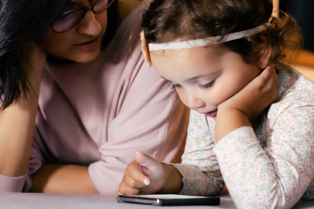 istock Small girl using smart phone while relaxing with mother at home. 1295101625