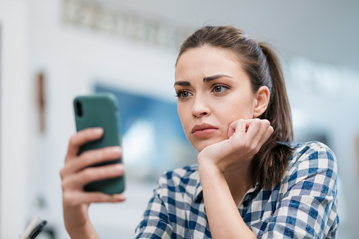 Close up of a depressed Caucasian young woman sitting at home, reading some bad news on her smart phone, feeling sad and worried