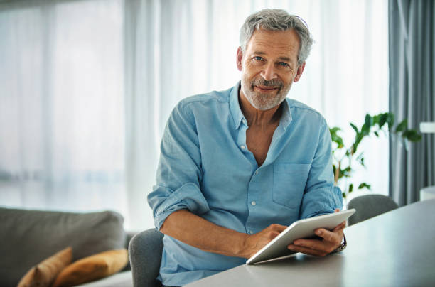 Mid aged man staying at home during coronavirus quarantine. Closeup side view of a mid 50's man staying at home during coronavirus quarantine. He's sitting in the living room as he's messaging his family members and/or looking for a job by using a digital tablet. Looking at the camera and smiling. mid adult stock pictures, royalty-free photos & images