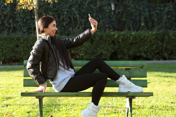 istock Young woman sitting on a bench in a public park using mobile phone for selfie 1295108078