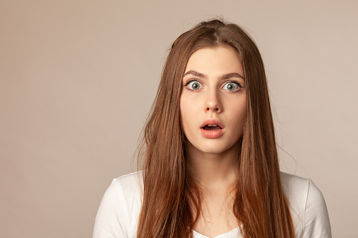 close up studio portrait of surprised 18 year old woman with long brown hair in white t-shirt on beige background