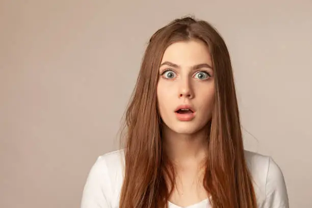 close up studio portrait of surprised 18 year old woman with long brown hair in white t-shirt on beige background