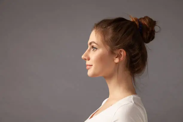 Close up studio portrait of attractive 18 year old woman with brown hair bun in white t-shirt on gray background