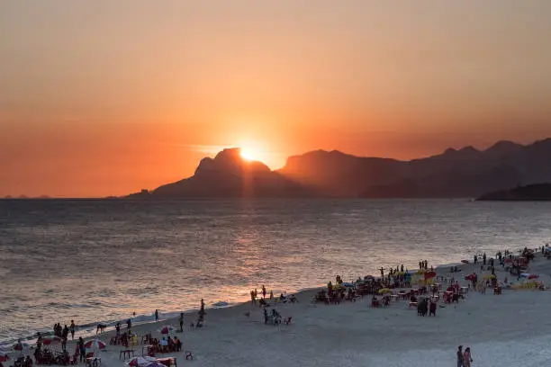 Orange sunset by the ocean in Piratininga, Niteri, with sun dipping behing the Gavea Stone in Rio de Janeiro. A beach full of people can be seen in the foreground in shadow.